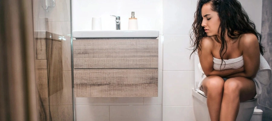 mulher sentada no vaso sanitário com as mãos na barriga e expressão de dor e desconforto abdominal