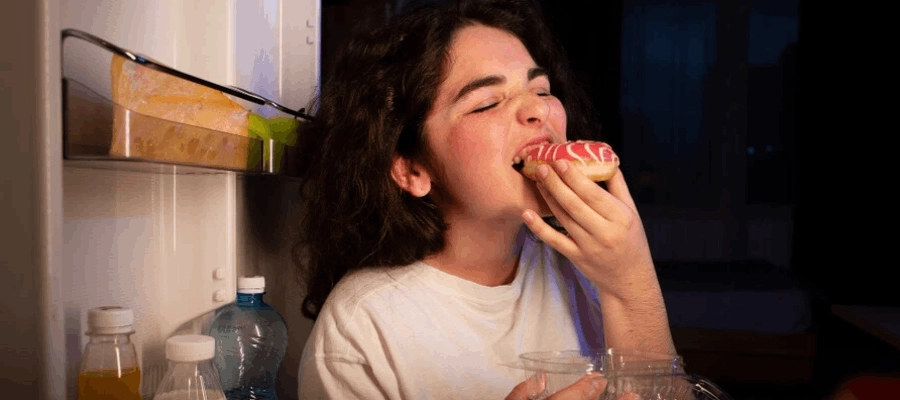 mulher jovem comendo em frente a geladeira porque ansiedade engorda