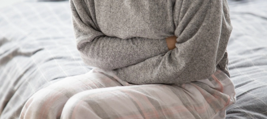 Mulher sentada na cama segurando o estômago com desconforto abdominal.